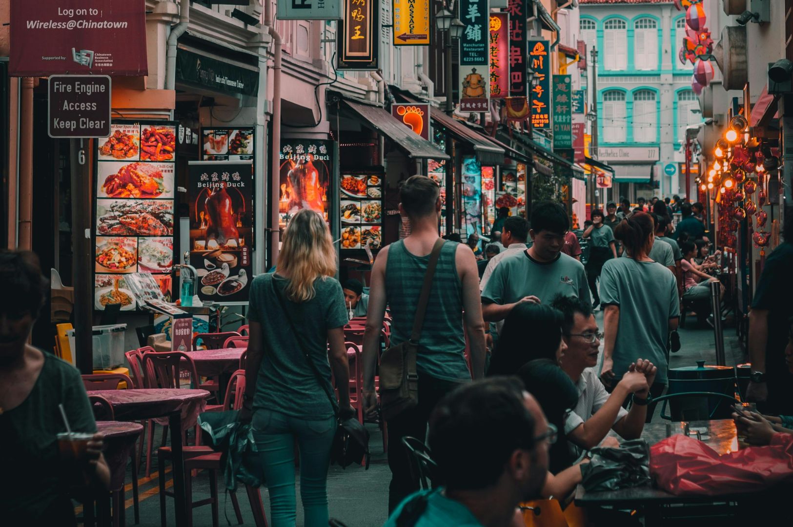 Busy street scene in Chinatown with vibrant signs and food displays. People walk and dine at outdoor tables, creating a lively, communal atmosphere.