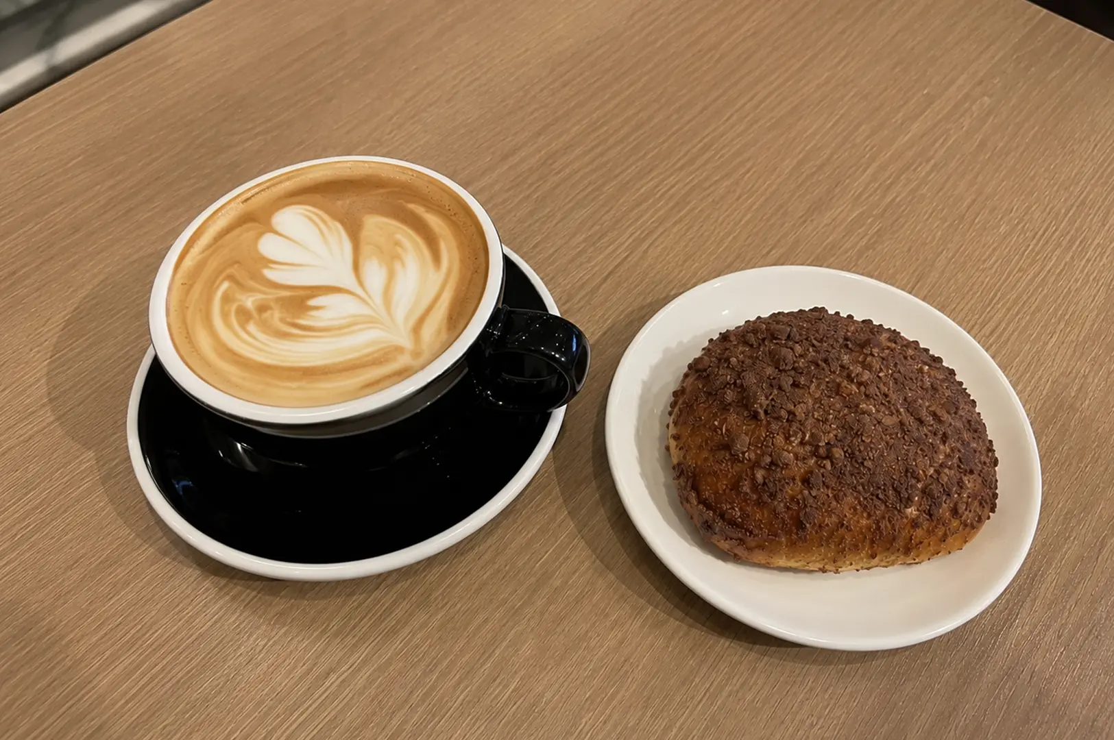 Latte with heart latte art served alongside a chocolate-topped sweet bun on a wooden table.