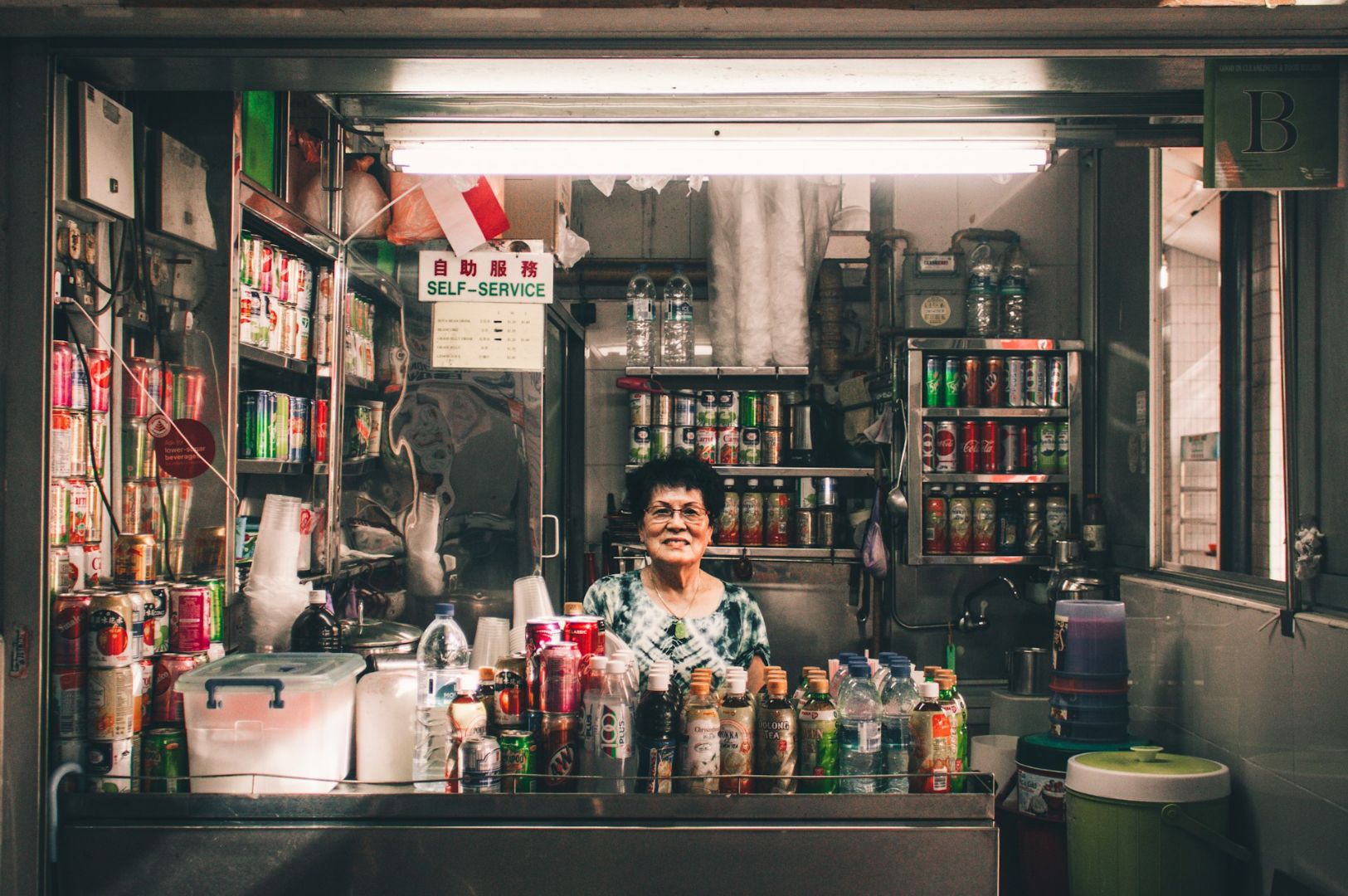 Elderly woman smiling behind a small shop counter filled with colorful drinks and canned goods. The sign above reads "Self-Service." Cozy and inviting.