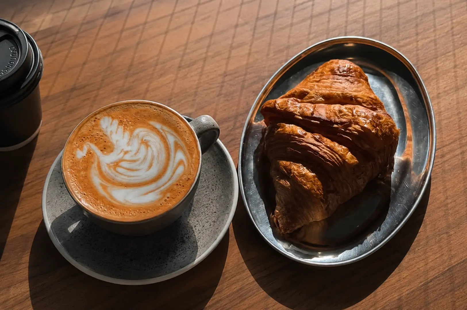 Fresh latte with latte art paired with a golden buttery croissant on a dark serving tray.