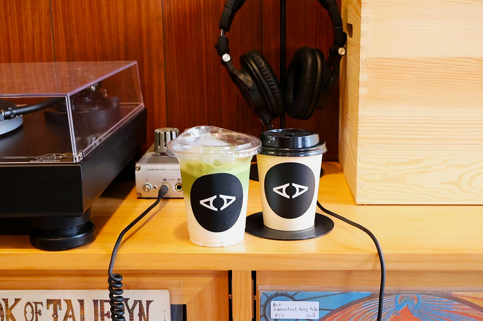Two drinks with a black and white logo sit on a wooden shelf. One is iced with a green hue, the other has a black lid. Headphones and a turntable are nearby.