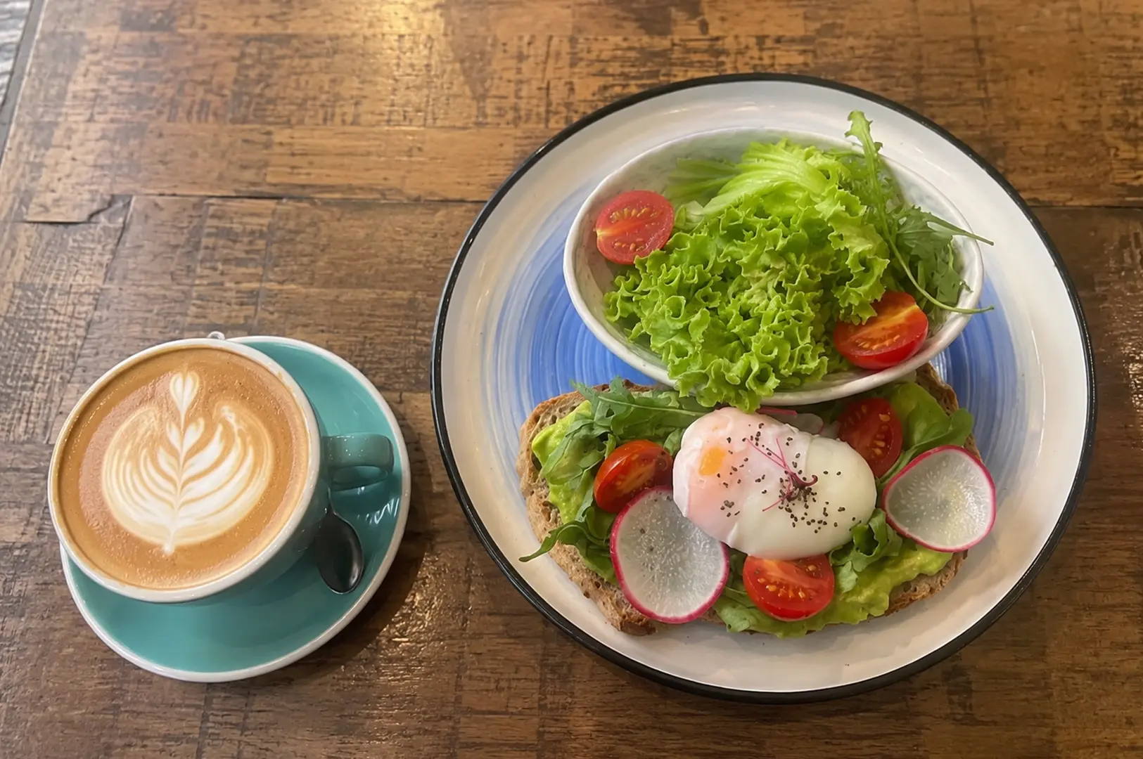 Avocado toast with poached egg, fresh side salad, and a latte on a rustic wooden table.