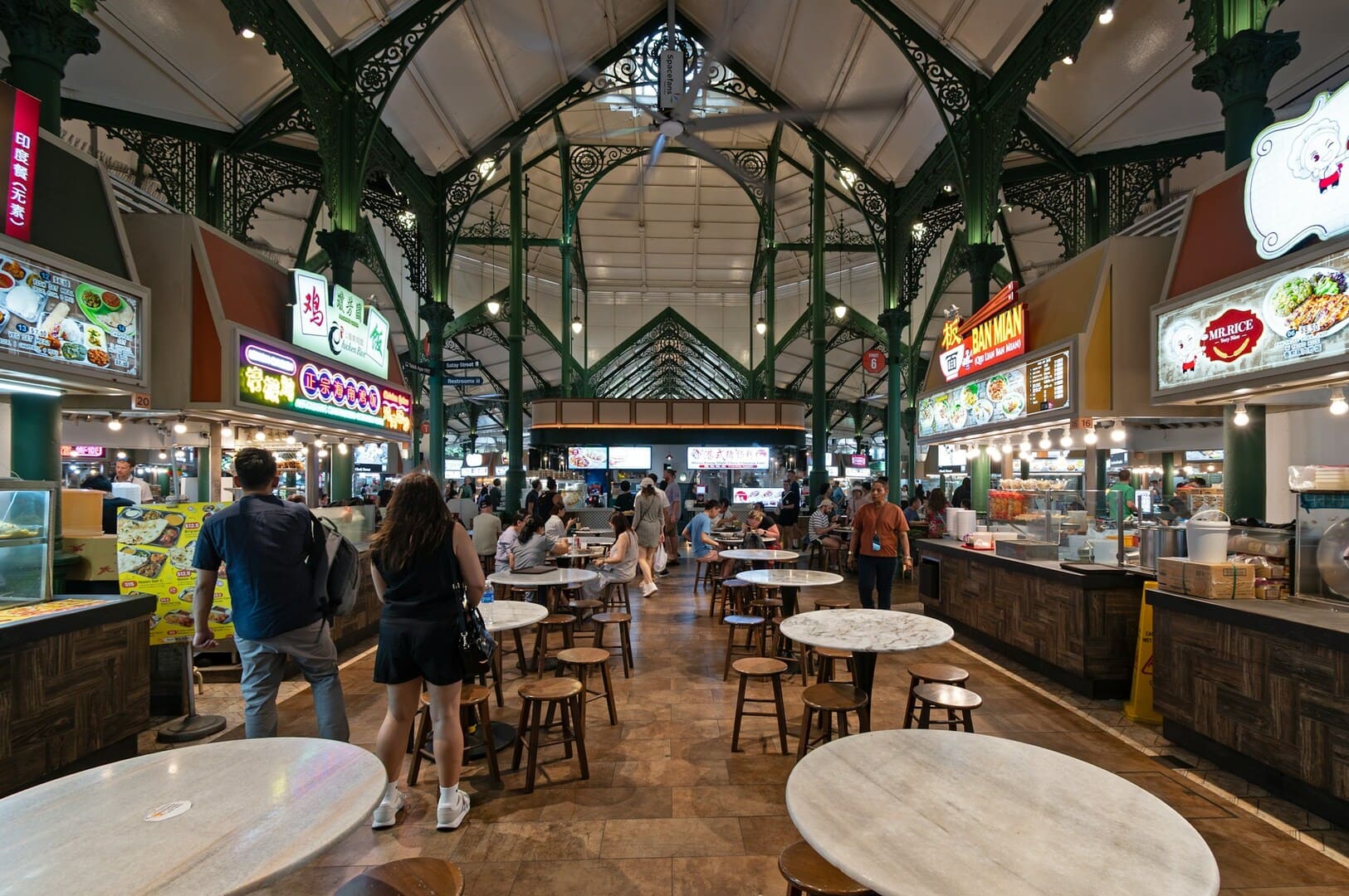 Bustling indoor food court with ornate green pillars; various food stalls line both sides. People walk and sit at round marble tables. Warm, lively atmosphere.
