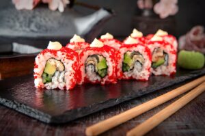 A black slate displays sushi rolls garnished with red sauce and cucumber, representing East Meets West Cuisine.