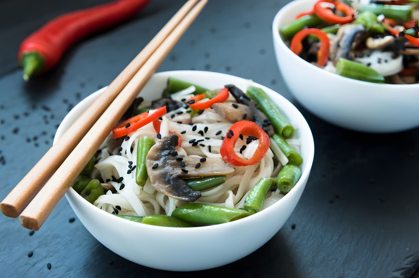 Two bowls filled with noodles and fresh vegetables on a sleek black table, showcasing East Meets West Cuisine.