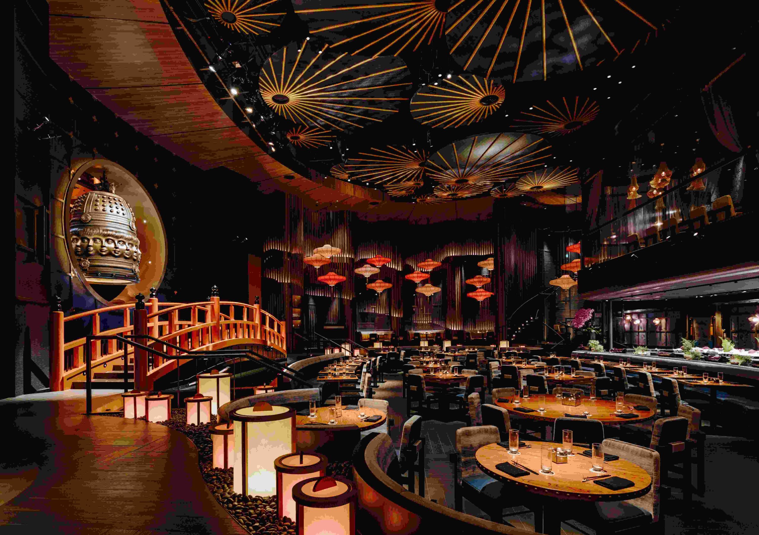 A luxurious and dramatic Japanese-inspired restaurant interior featuring a ceiling decorated with golden umbrellas, a wooden bridge, and a large ornamental bell.
