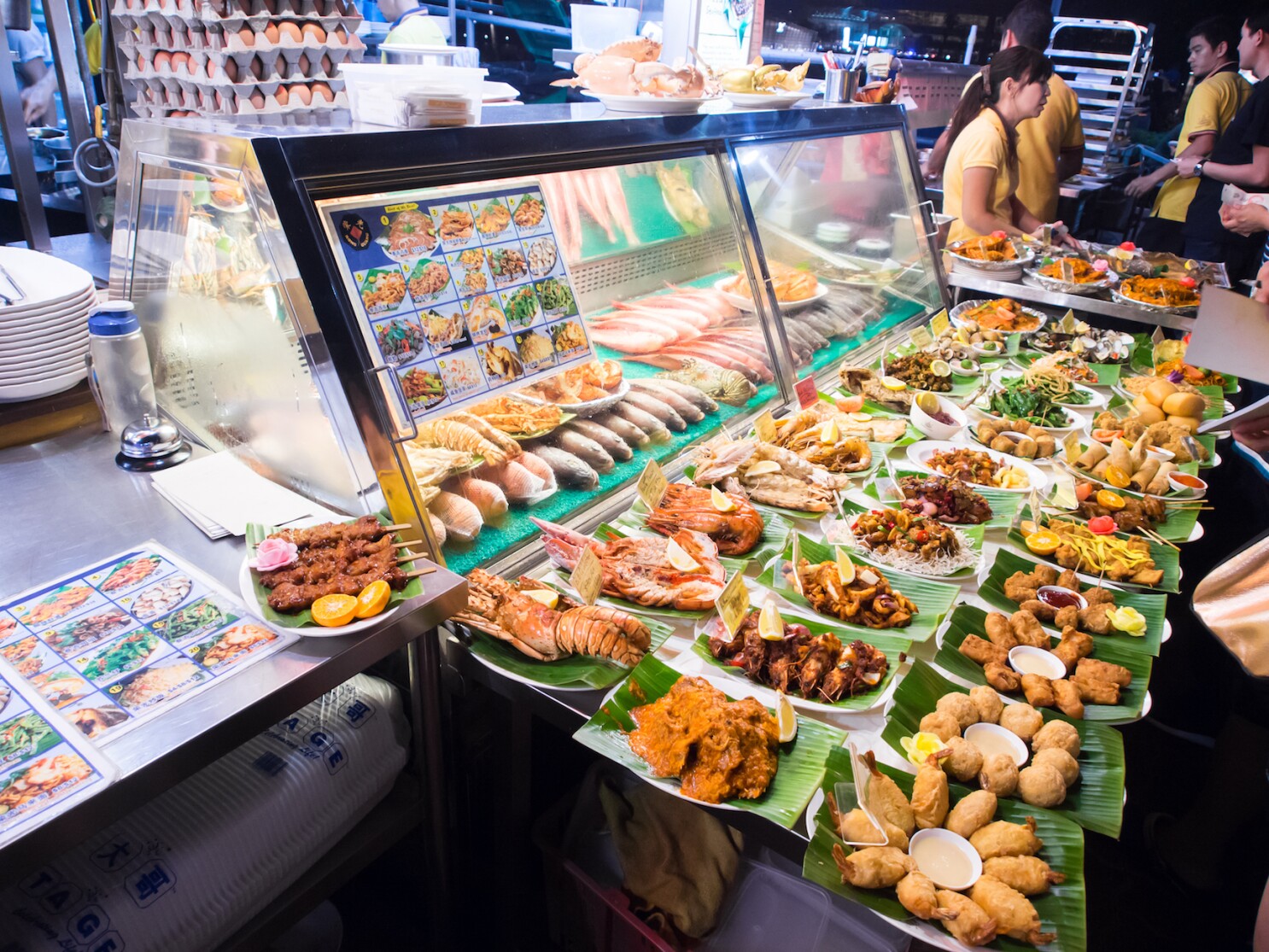 A colorful display of prepared Singaporean hawker food, including fresh seafood, prawns, satay skewers, and crispy dishes on banana leaves at a street food stall.