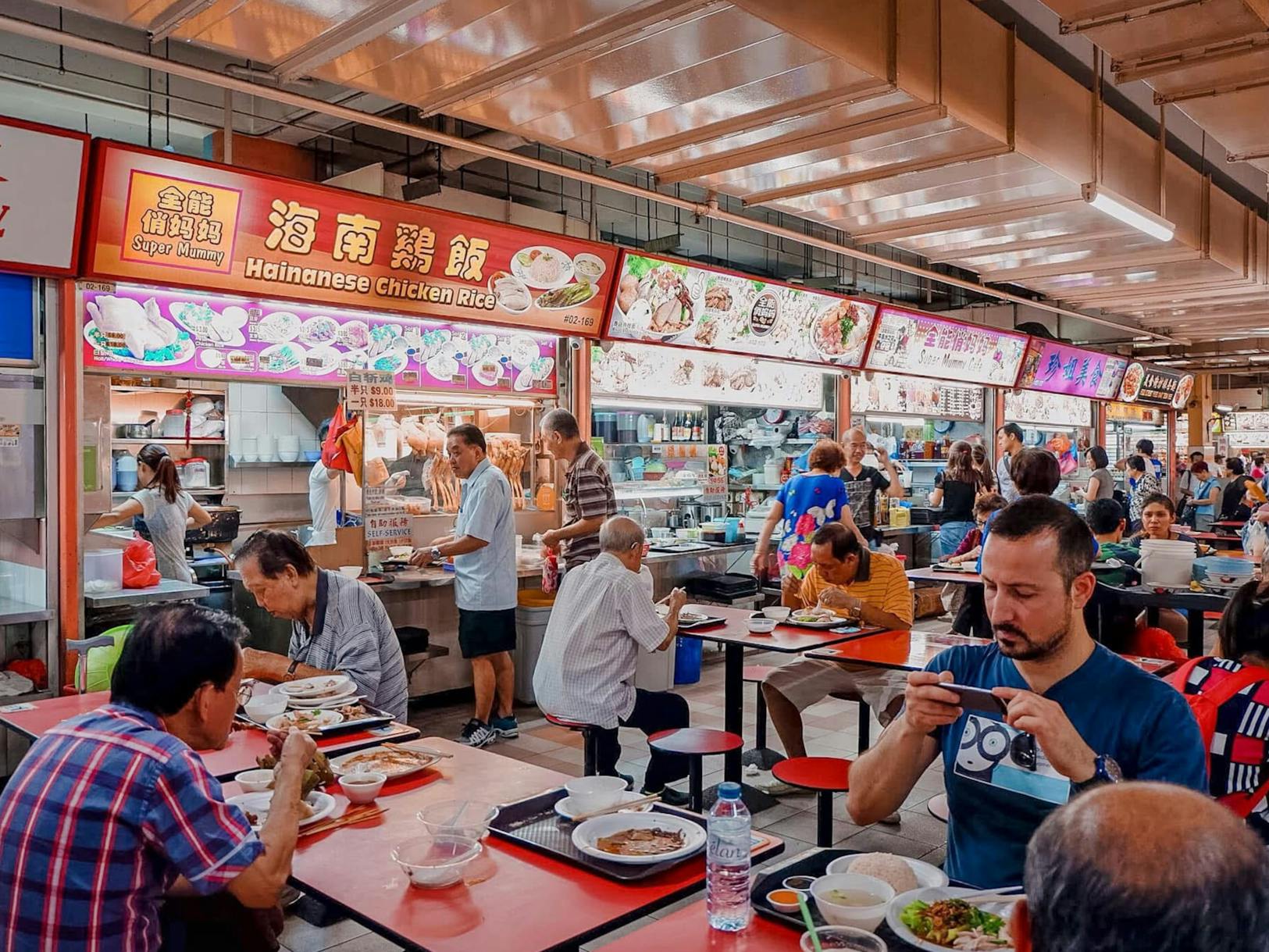 A busy Singapore hawker center with patrons enjoying Hainanese Chicken Rice and other local cuisine at red tables, showcasing the authentic and vibrant street food culture.