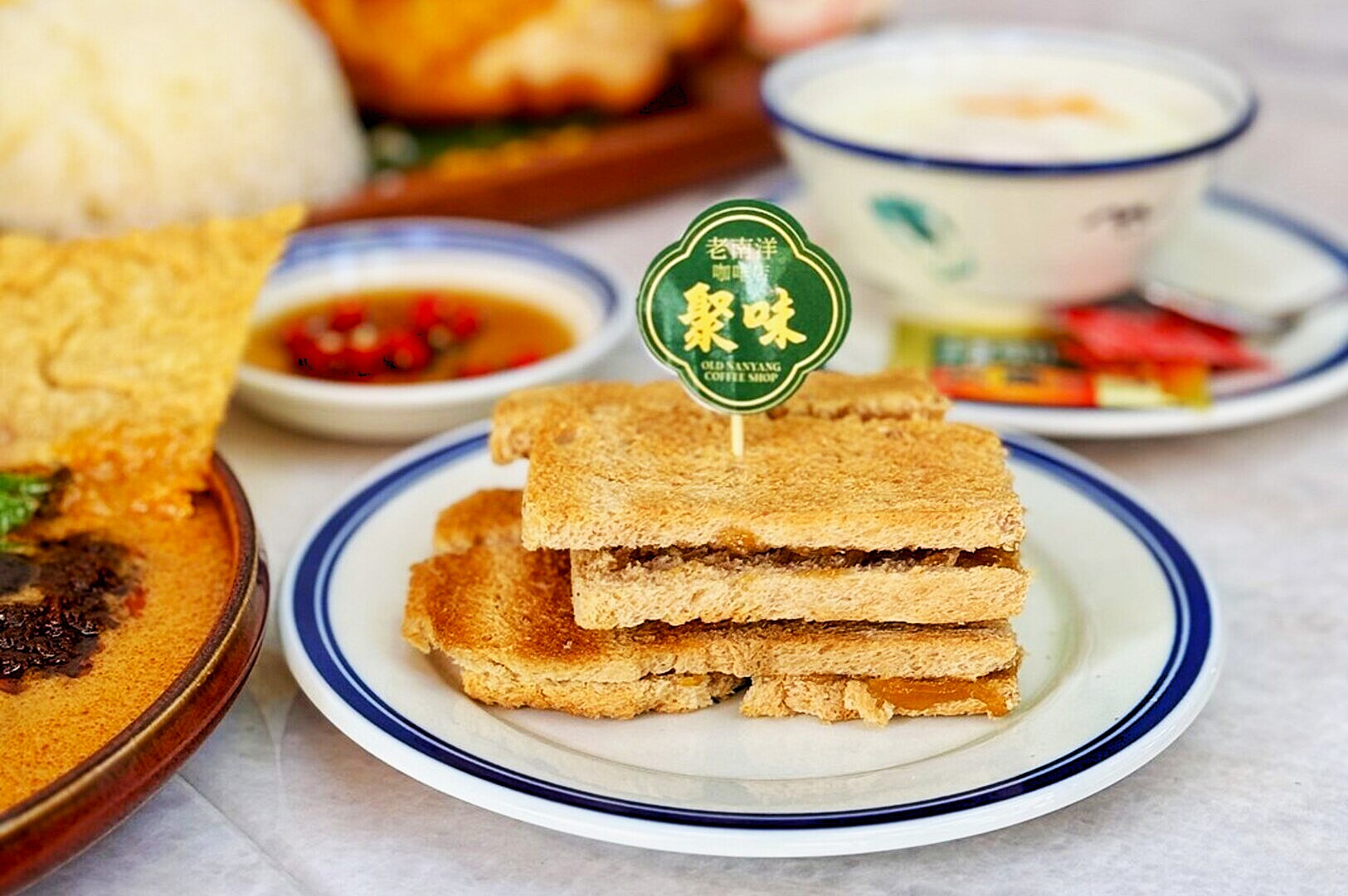 Toasted kaya sandwiches on a white plate with a toothpick sign. Background shows a bowl of soup, a dish with rice, and a small sauce plate.