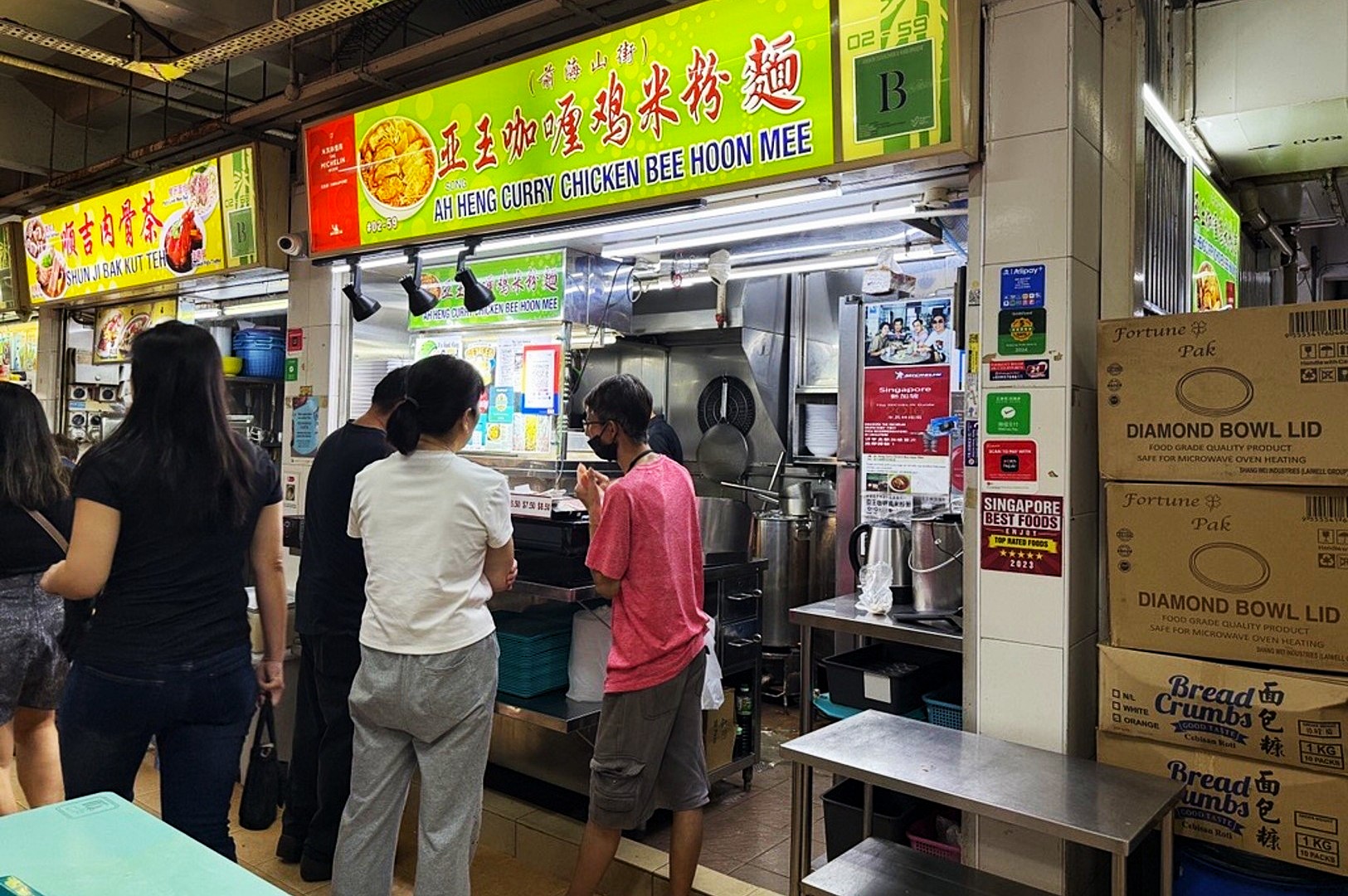 People waiting in line at a bustling food stall named "Ah Heng Curry Chicken Bee Hoon Mee" in a hawker center. Bright signage above, and boxes stacked nearby.