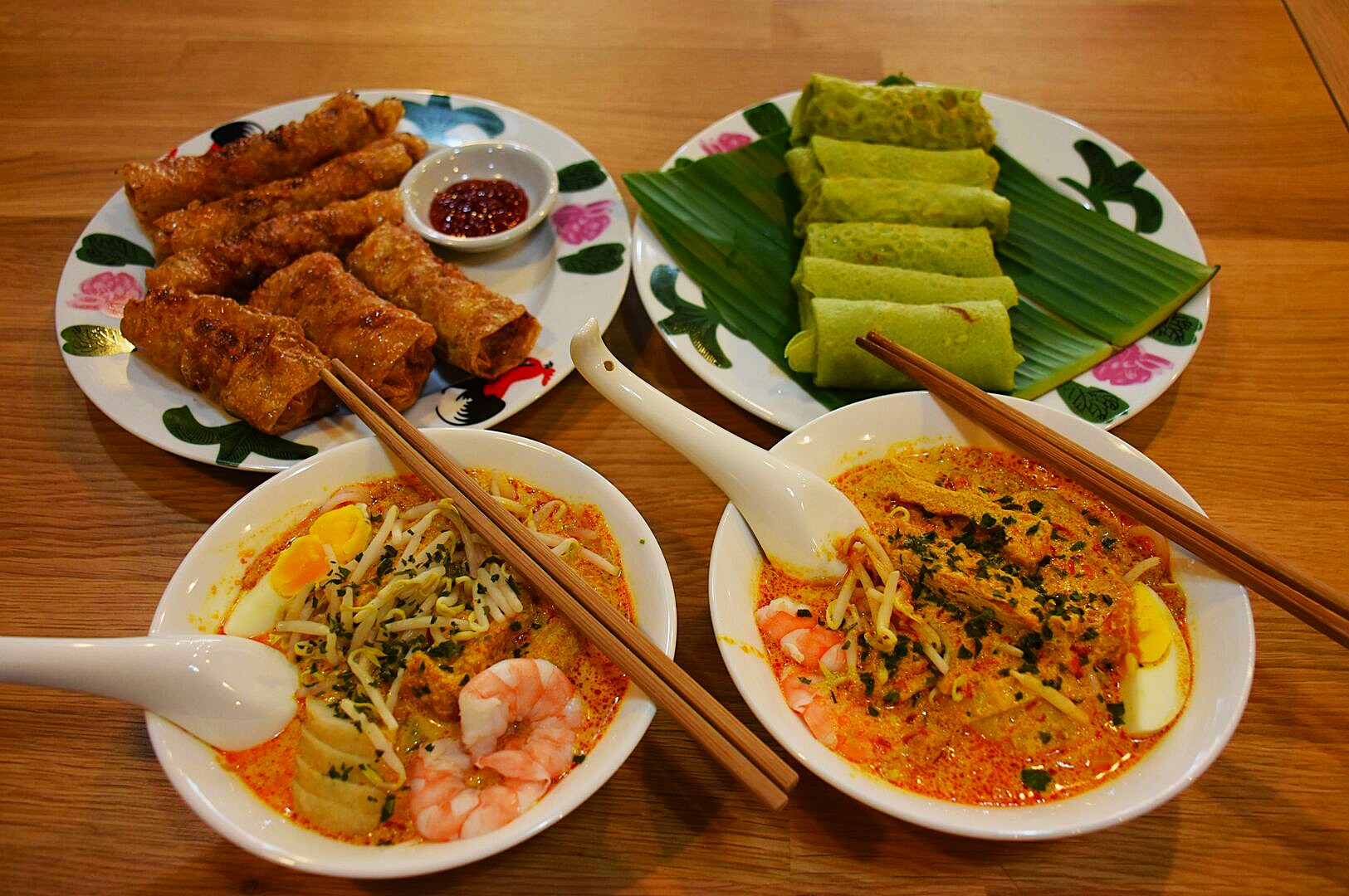 A vibrant meal featuring two bowls of shrimp and egg laksa, a plate of crispy spring rolls with dipping sauce, and green pancakes on banana leaves.