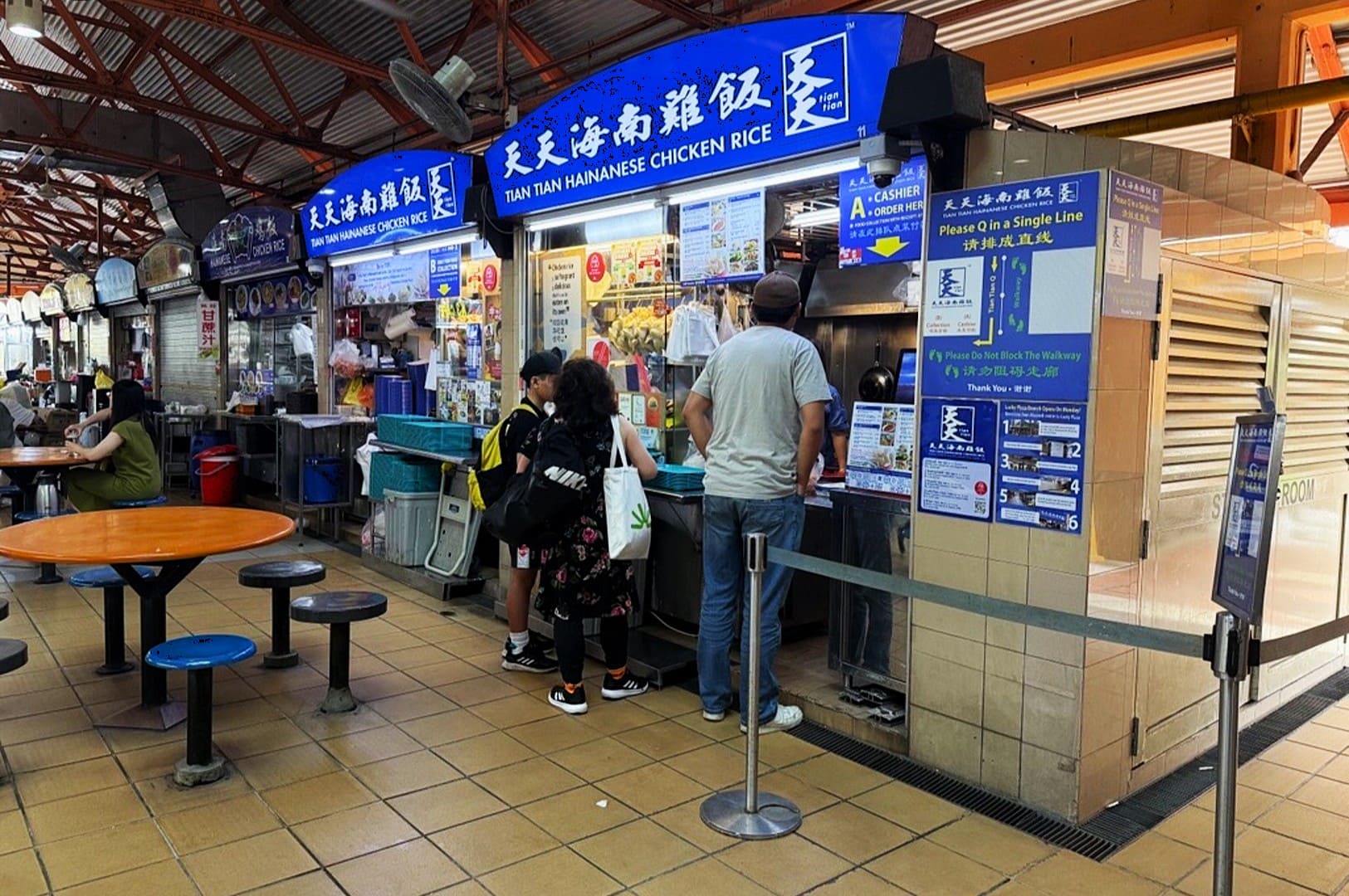 People wait in line at a food stall named "Tian Tian Hainanese Chicken Rice" inside a bustling hawker center. Tables are nearby, and the atmosphere is lively.