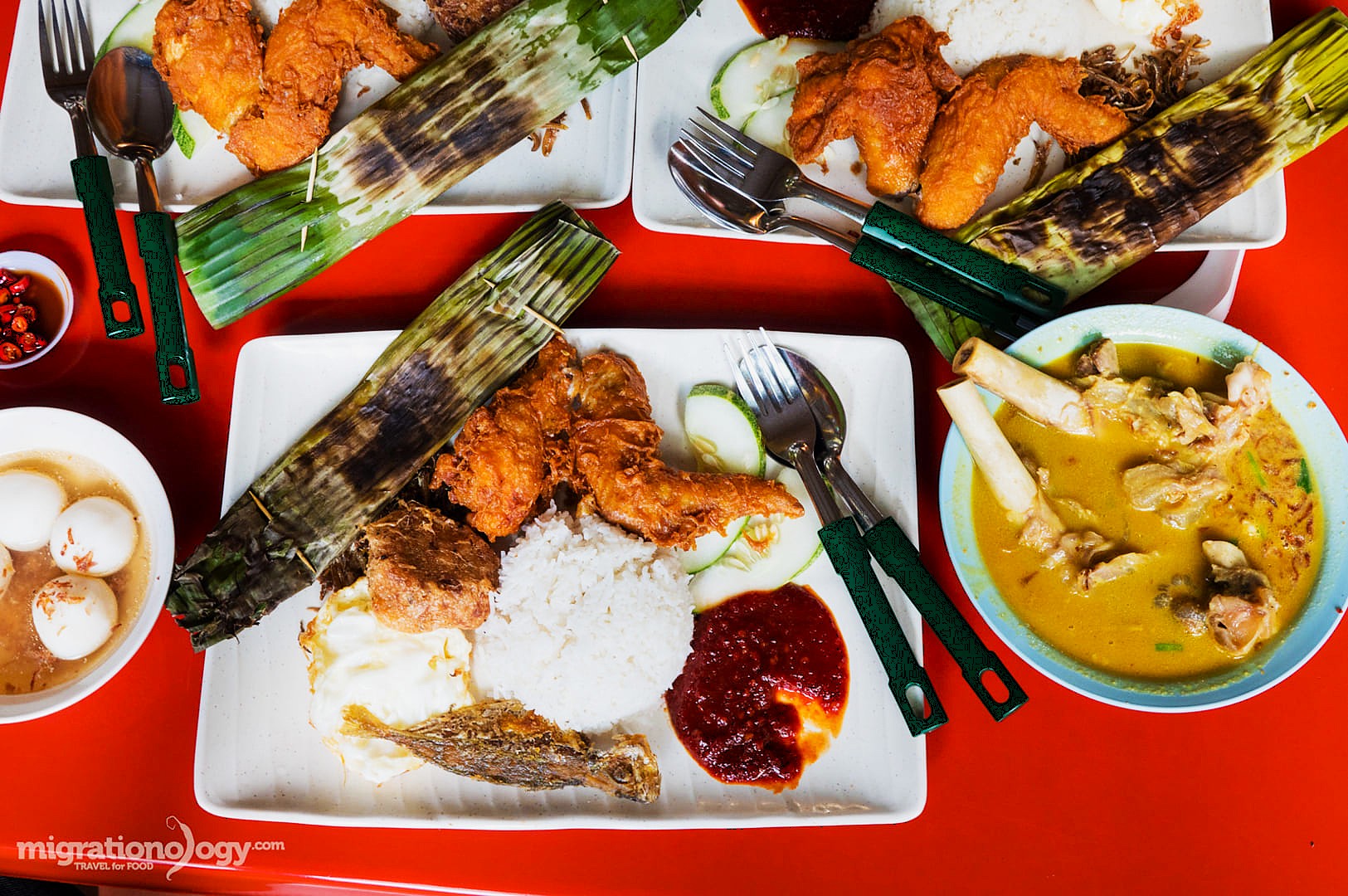 Plates of Malaysian nasi lemak with fried chicken, rice, sambal, and cucumber slices. A bowl of yellow curry soup and hard-boiled eggs are on the side.