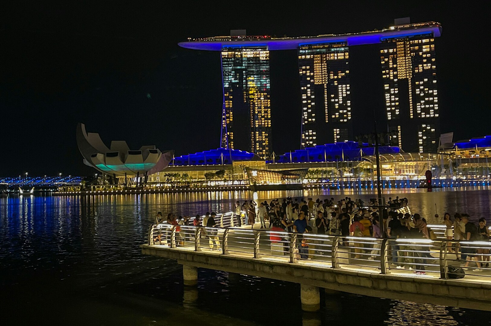 Nighttime scene of people on a dock beside a large building, the Waterfront Dining Restaurant at Marina Bay & Collyer Quay.