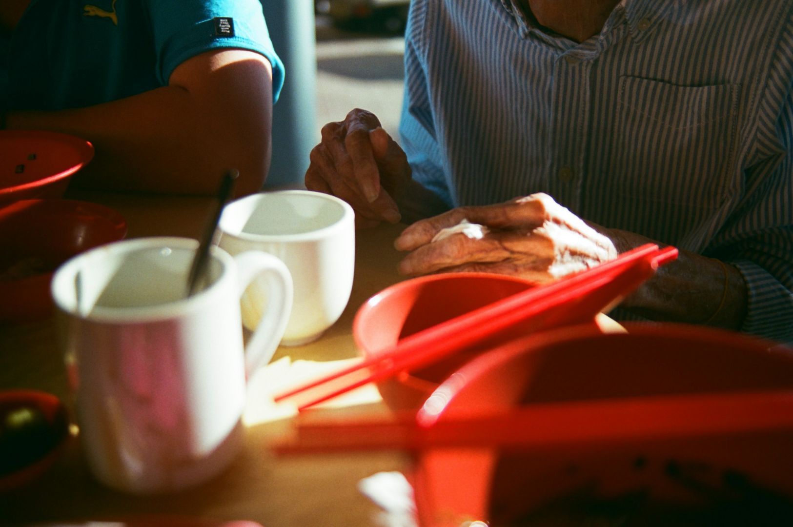 Close-up of a table with red bowls, red chopsticks, and white mugs. A person in a striped shirt with elderly hands is gently touching the table, suggesting a peaceful, domestic setting. Bright sunlight casts shadows.