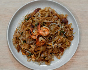 A plate of char kway teow stir-fried with flat rice noodles, prawns, egg, and bean sprouts, glistening with dark soy sauce and served hot in a hawker-style presentation.