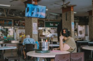 A woman sits alone at a table in a food court, watching a TV screen above. The scene is relaxed, with empty chairs and a counter visible in the background.