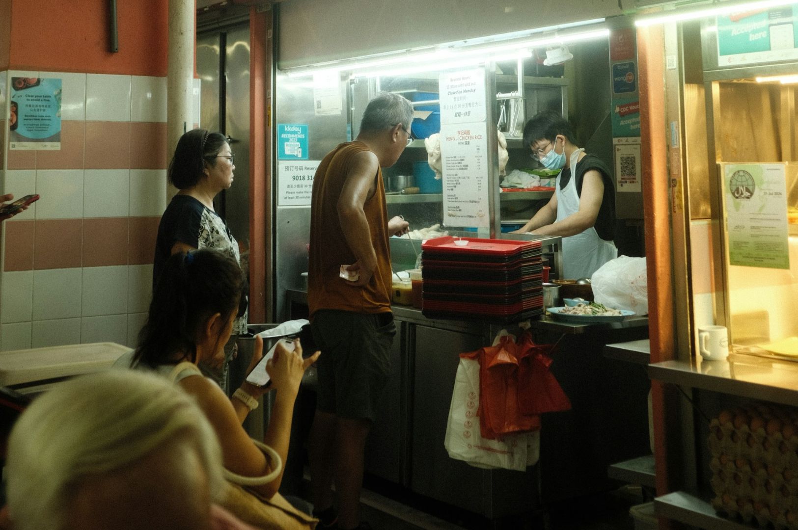 People line up at a food stall. A vendor in a mask prepares meals, while customers, including a seated woman, wait. Warm, bustling atmosphere.
