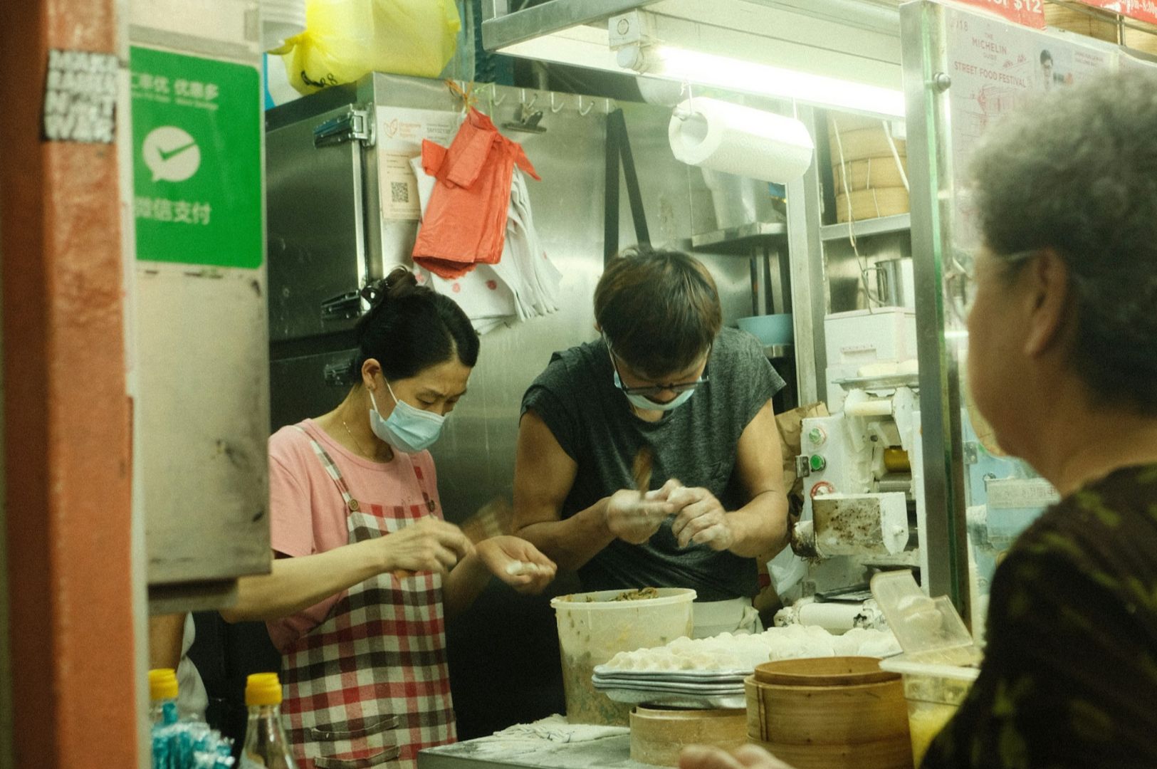 A bustling kitchen scene shows two masked cooks, a man and a woman, preparing dumplings. An elderly customer observes, creating a lively atmosphere.