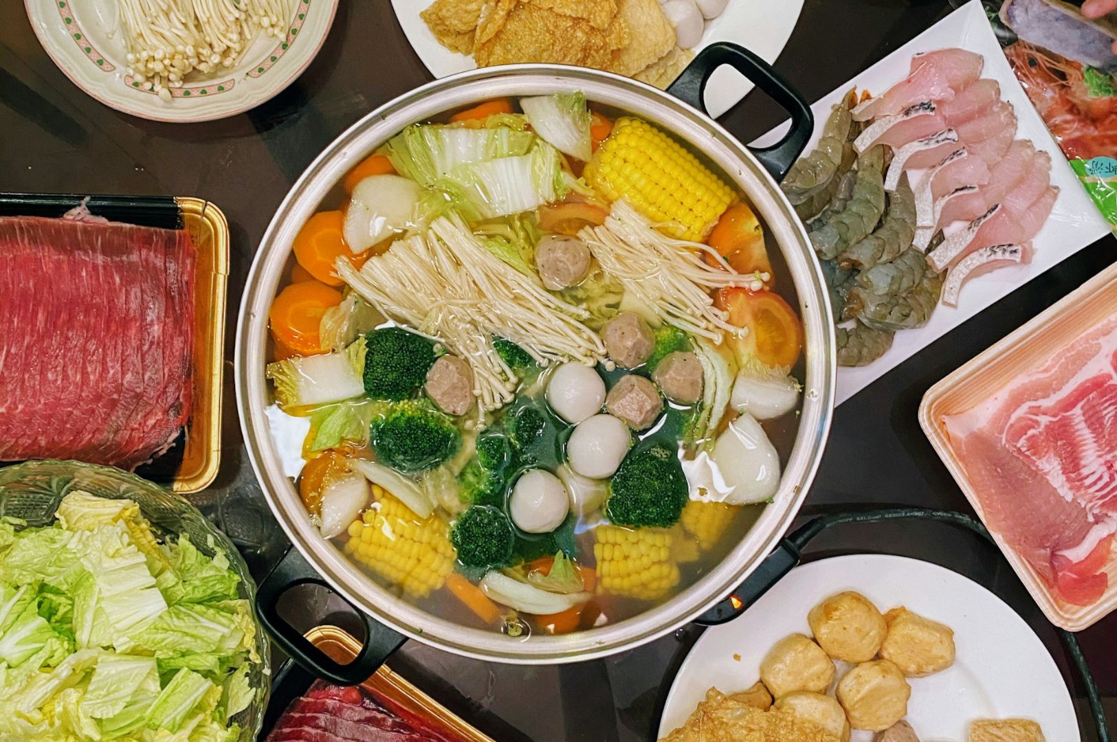 A colorful hot pot filled with vegetables, mushrooms, corn, and meatballs. Surrounding plates hold sliced meats, seafood, and more vegetables.
