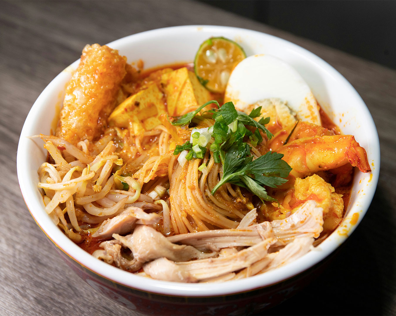 A steaming bowl of laksa with rich coconut-based broth, topped with prawns, fish cake, and fresh herbs, served in a deep ceramic bowl.