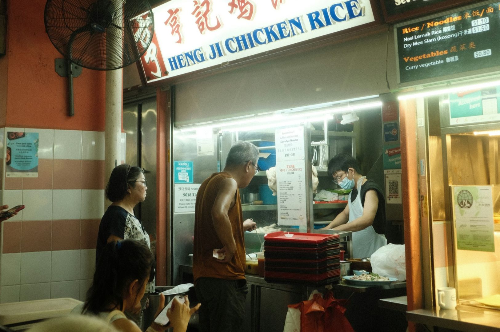 A hawker stall with a sign reading "Heng Ji Chicken Rice." A masked vendor serves dishes, while two customers wait their turn. The setting is busy and casual.