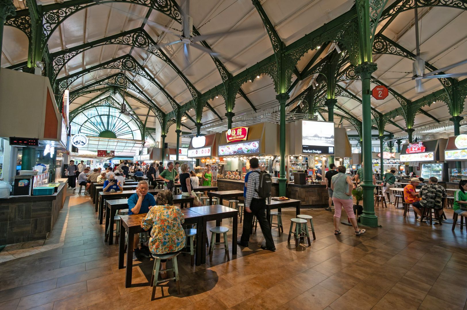 A bustling food hall with ornate green iron pillars and high ceilings. People sit at wooden tables, enjoying meals, while others explore various food stalls.