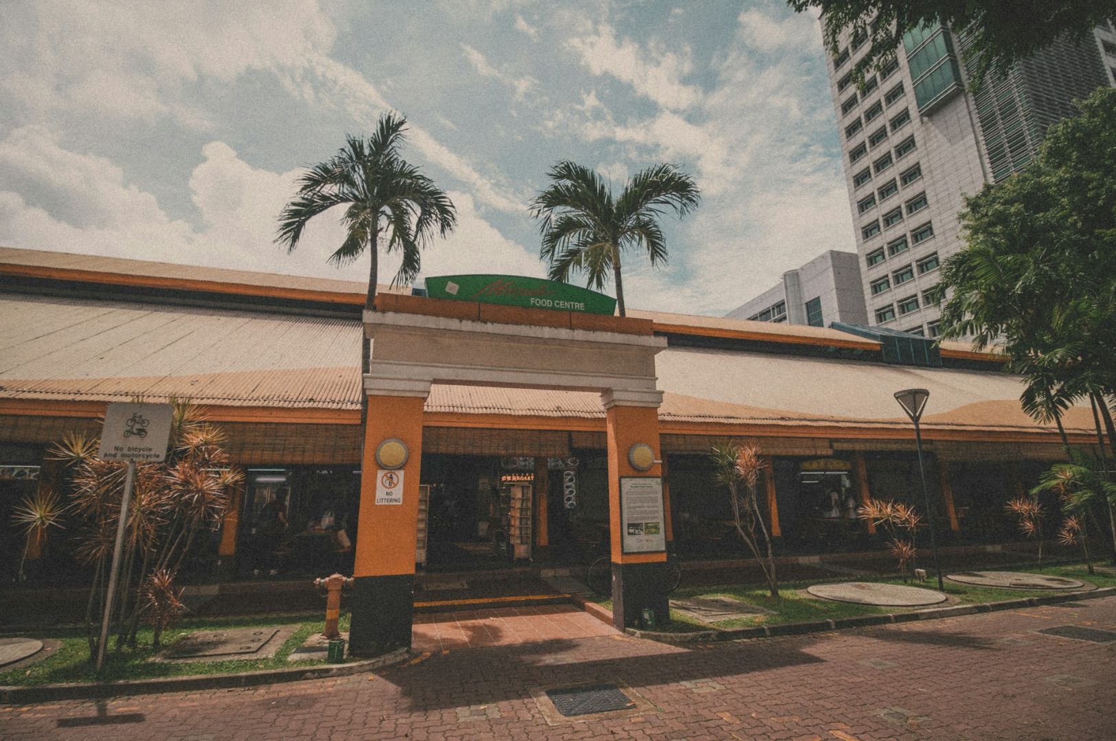 A large food center with tropical palm trees at the entrance is under a partly cloudy sky. The atmosphere is calm and inviting.