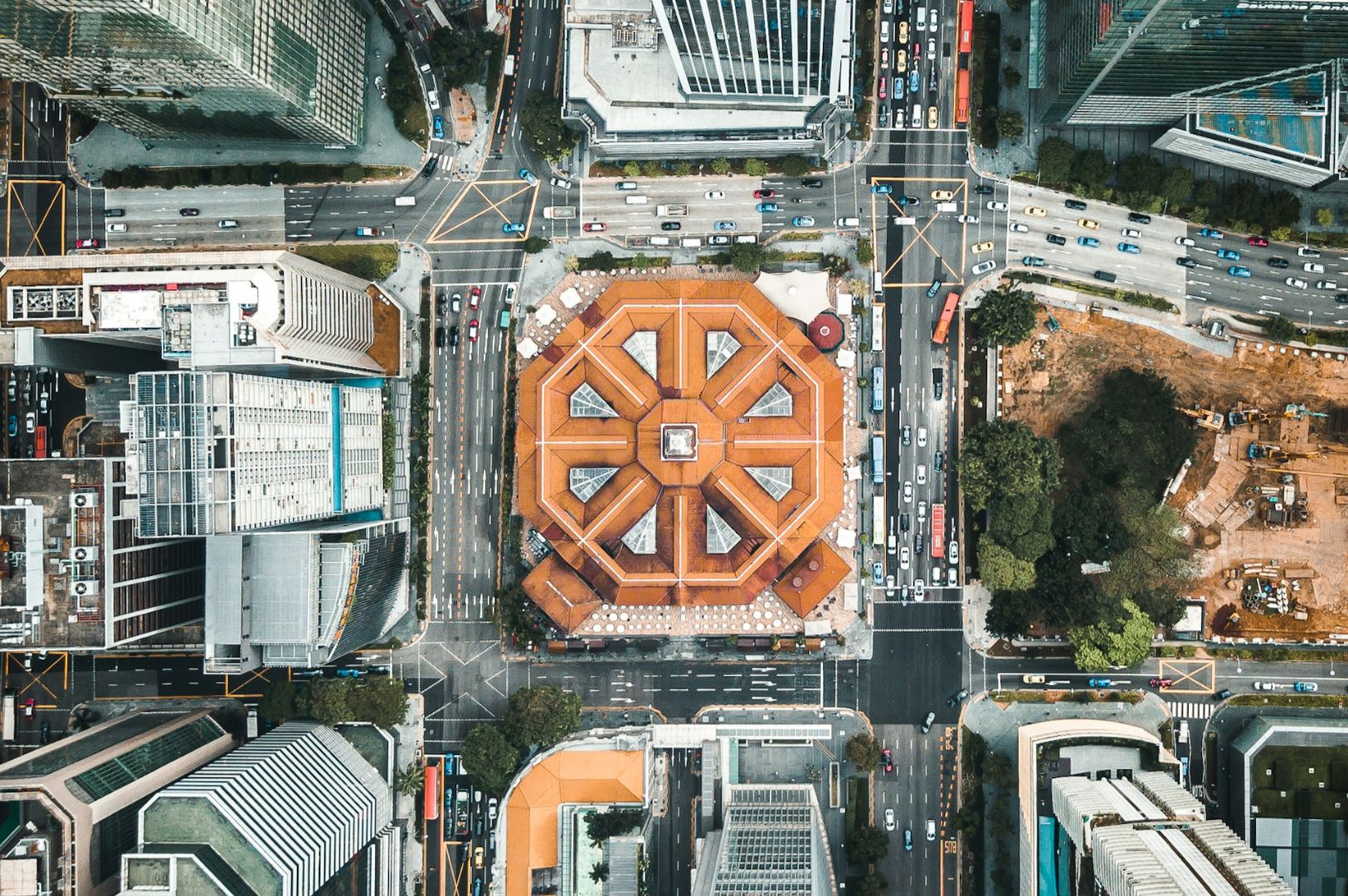 Aerial view of a busy urban intersection with a striking orange-roofed, octagonal building at the center. Surrounding are modern skyscrapers and a construction site.