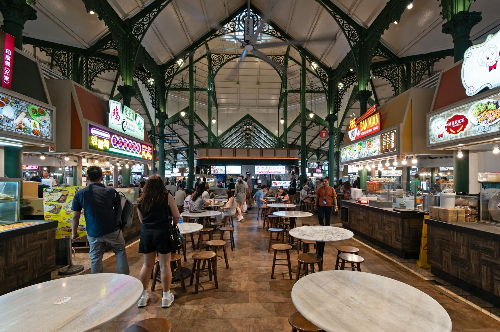 Spacious food court under a high, ornate ceiling with various food stalls. People walk and sit at round tables, creating a lively, bustling atmosphere.