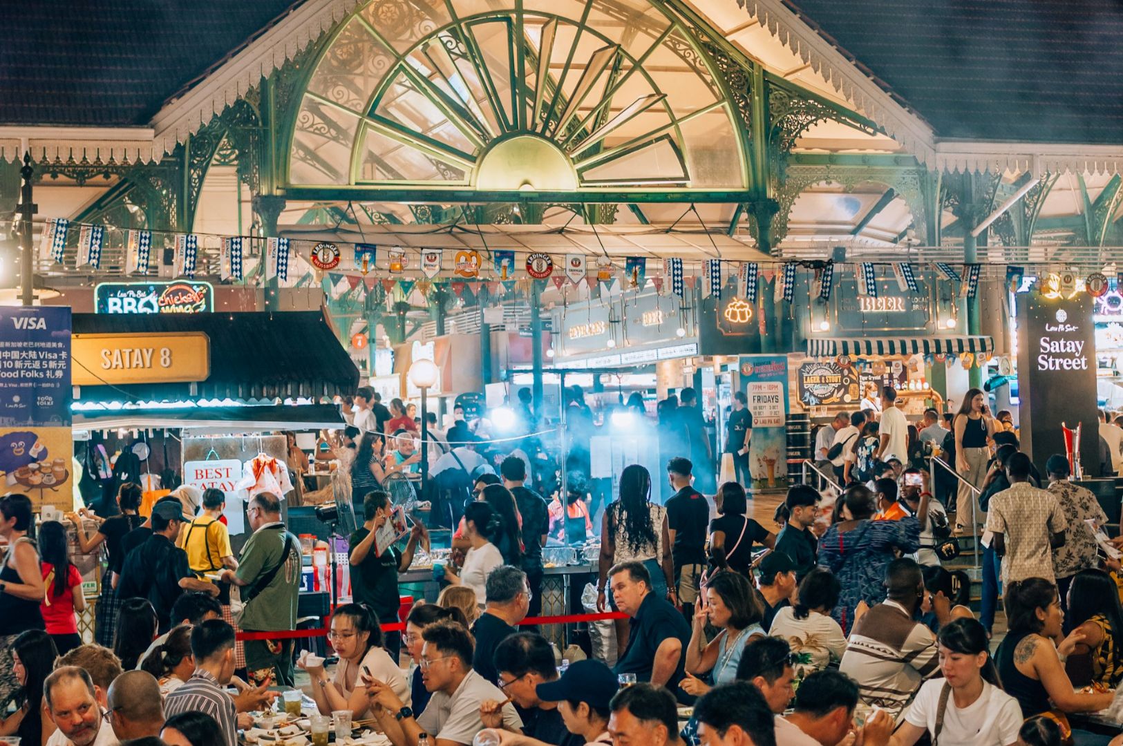 Bustling food market with diverse crowd enjoying meals. Stalls display signs under ornate structure, lively atmosphere, smoke from grilled food.