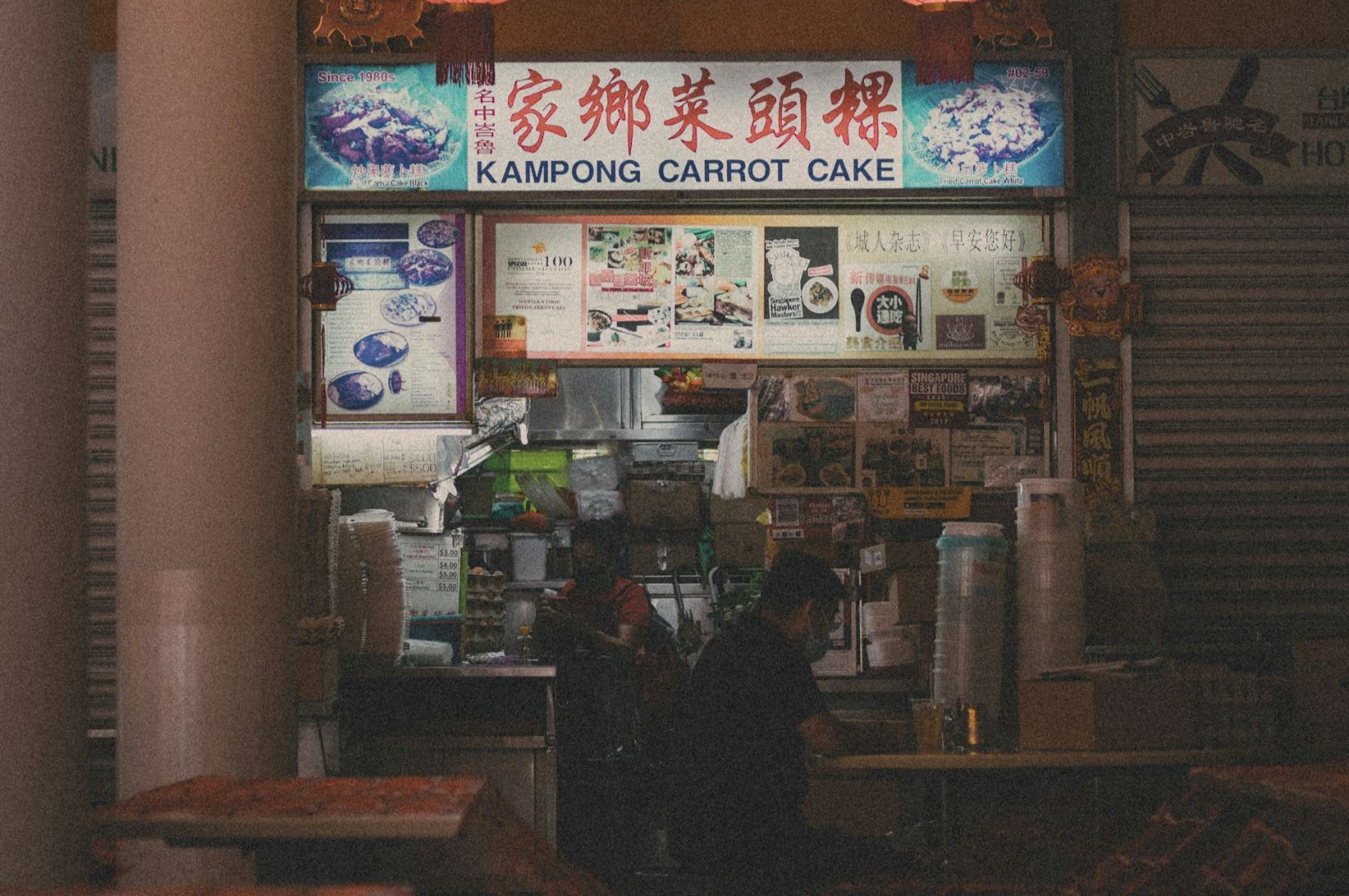 A dimly lit food stall with a "Kampong Carrot Cake" sign in a vibrant market. Menu and colorful pictures adorn the stall, creating a cozy, inviting atmosphere.