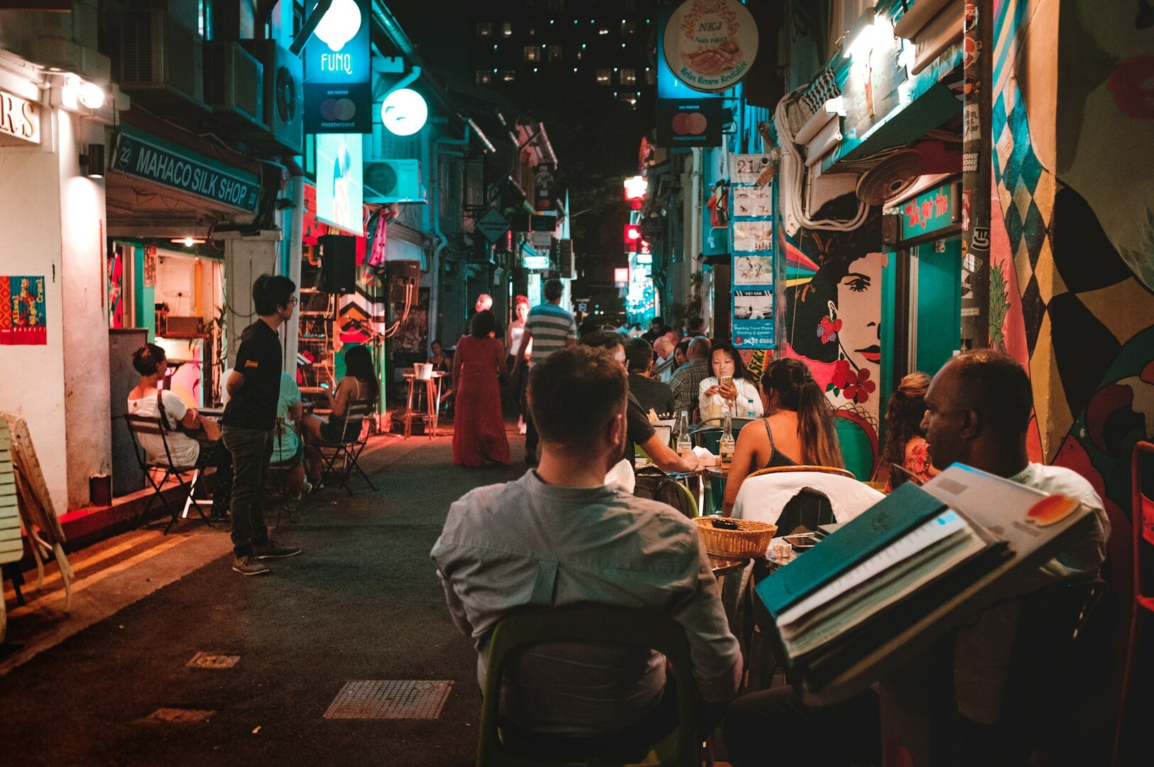 A vibrant, bustling alley at night, with people dining at tables under colorful lights. Walls are adorned with murals, creating a lively, urban atmosphere.