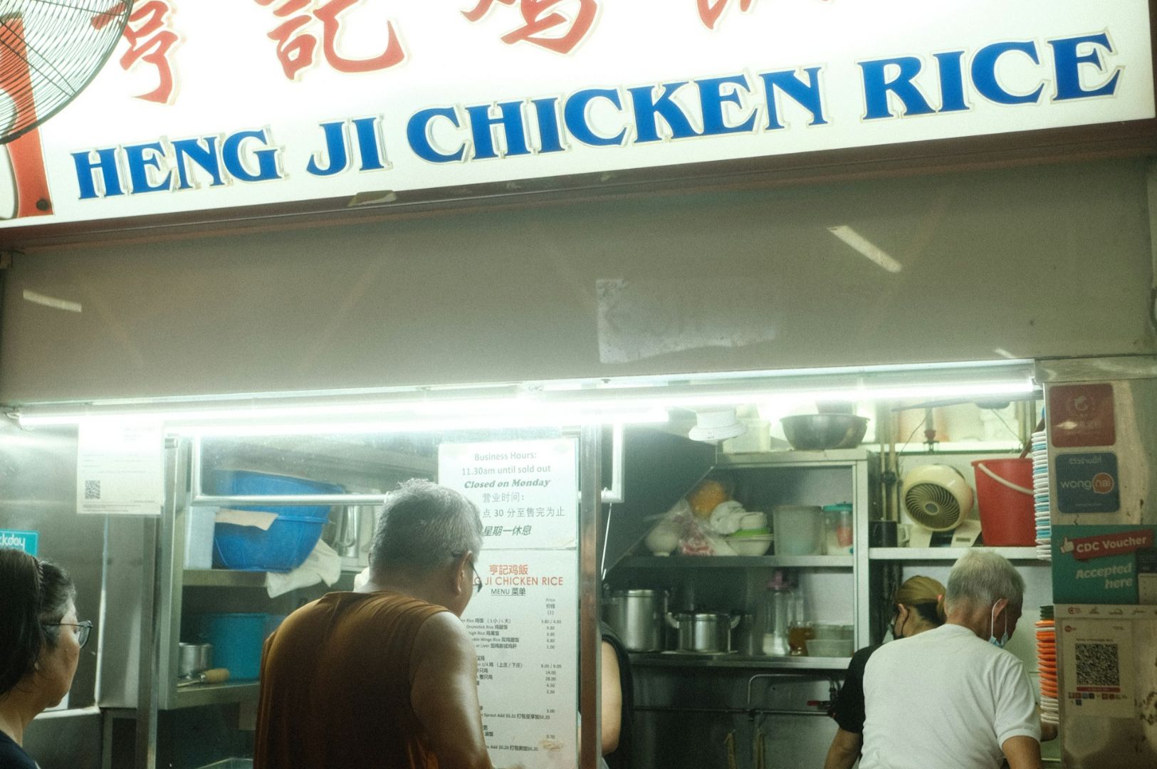 People stand in front of a bustling chicken rice stall named "Heng Ji Chicken Rice." The atmosphere is lively, and the shop appears busy.
