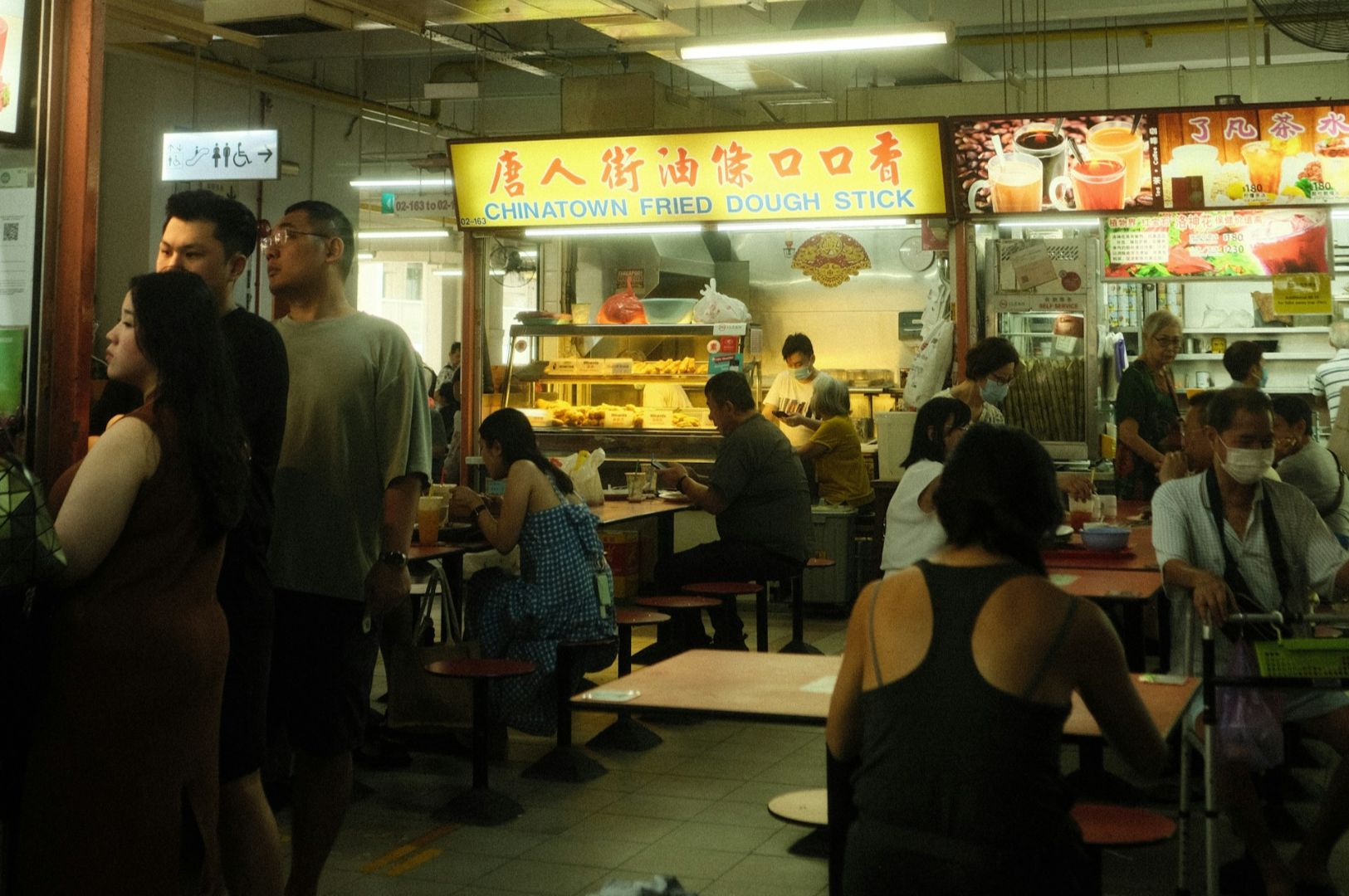 Busy indoor food court with a stall named "Chinatown Fried Dough Stick." People are eating and socializing, creating a lively atmosphere. Dim lighting.