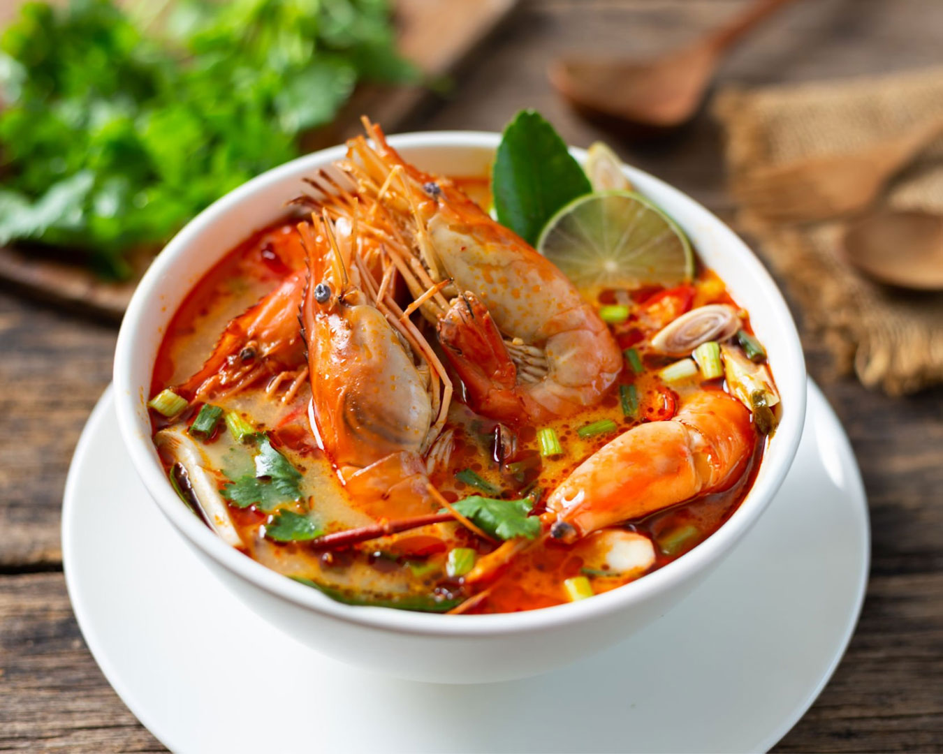 A bowl of spicy and sour tomyam soup filled with prawns, squid, and mushrooms, with fresh herbs floating in the clear reddish broth.
