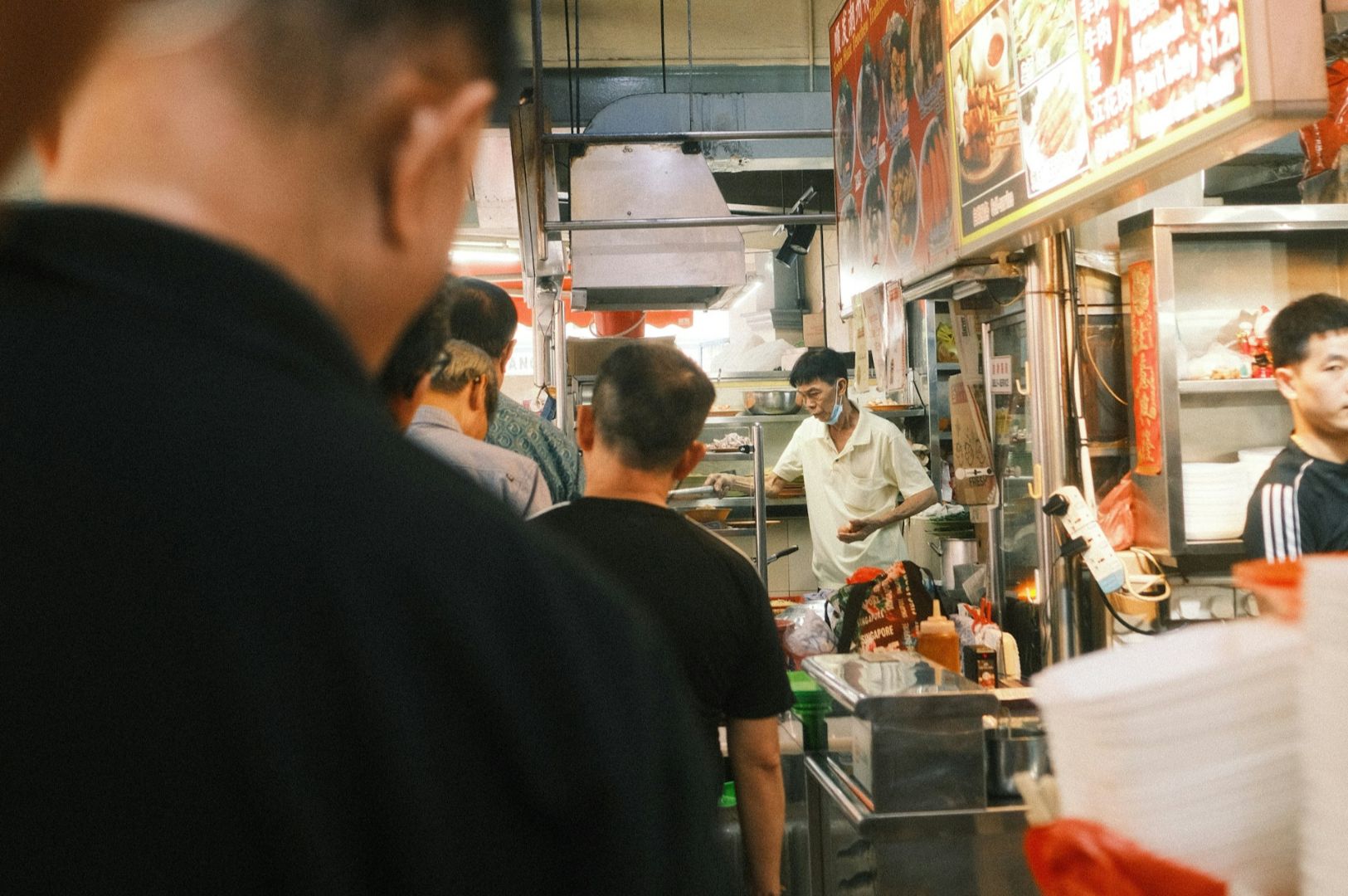 Indoor food court with people in line at a vendor's stall. Bright menu signs above, vendor preparing food, bustling atmosphere, warm lighting.