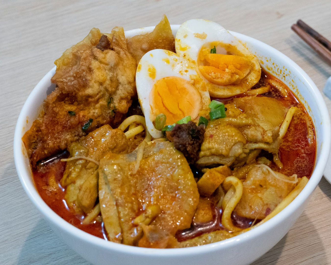 A bowl of curry noodles in creamy spiced broth, topped with tofu, prawns, and long beans, served hot with chilli paste on the side.