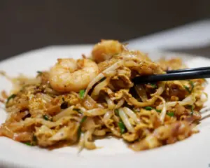 A close-up of a freshly plated Char Kway Teow, featuring glossy flat rice noodles stir-fried with prawns, cockles, eggs, and bean sprouts, garnished with chives