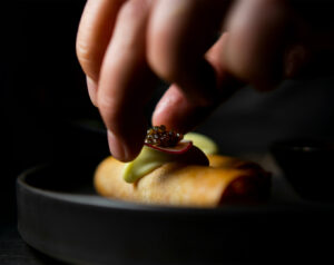 A chef’s hand gently lowers a plated dish onto the table, capturing the precise moment of presentation with soft lighting that highlights the textures and colours of the food.
