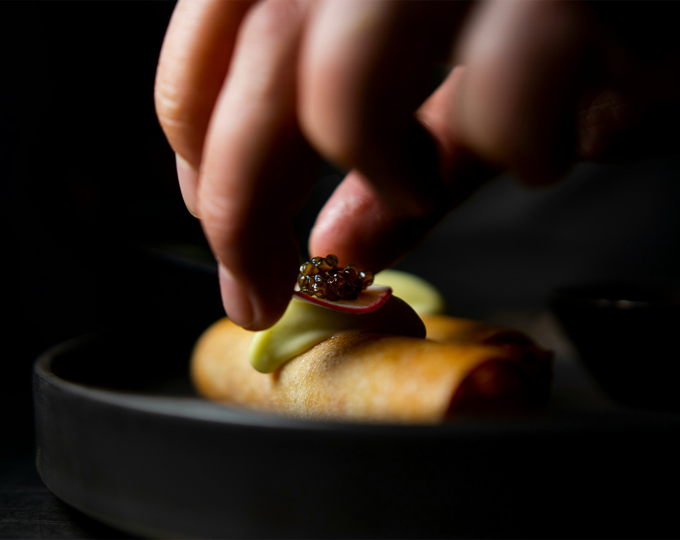 A chef’s hand gently lowers a plated dish onto the table, capturing the precise moment of presentation with soft lighting that highlights the textures and colours of the food.