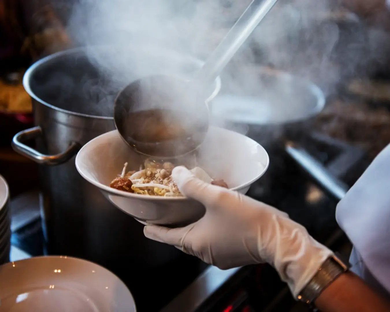 A hand gently pouring hot soup into a bowl of noodles, steam rising as the broth fills the bowl, highlighting the preparation of a comforting noodle dish.