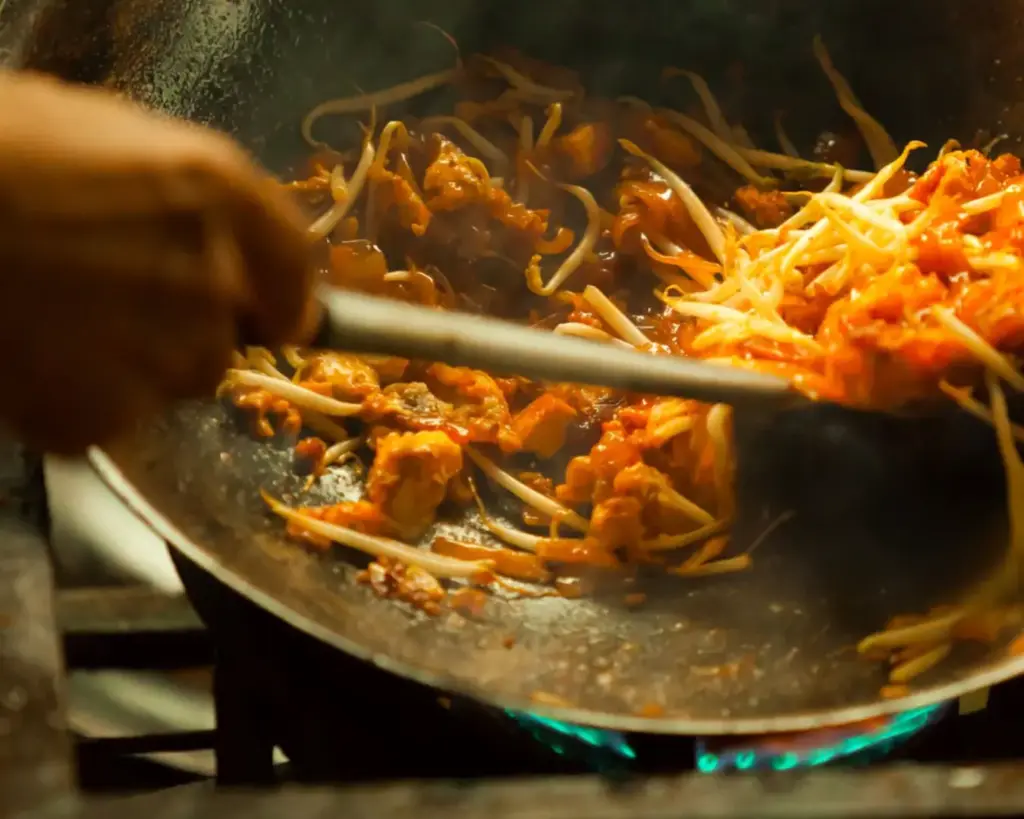 A street food vendor stir-frying Char Kway Teow in a large, well-seasoned wok over high heat. Flames rise beneath the wok as the noodles are tossed together with prawns, eggs, and dark soy sauce, creating the signature smoky aroma known as “wok hei.