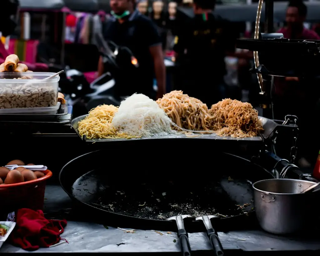 A bustling street food stall specialising in Char Kway Teow. The setup includes stacks of plates, fresh ingredients on display, and a lively atmosphere