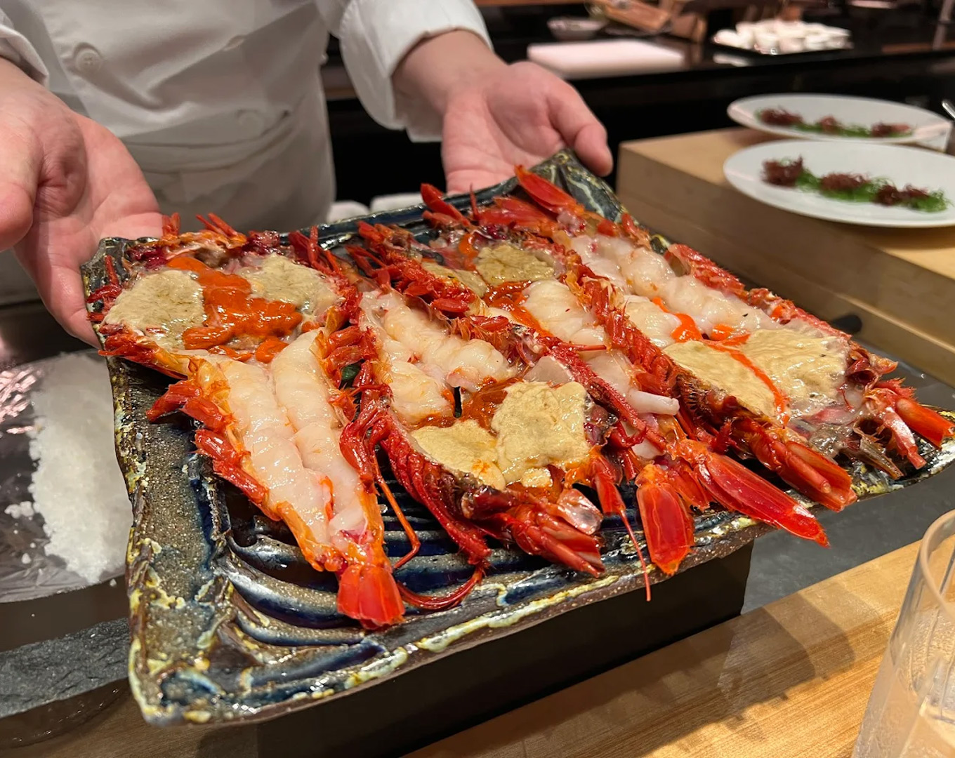 A hand presents a dish of marinated Botan shrimp topped with fresh sea urchin, showcasing translucent textures and a luxurious pairing of ocean flavours.