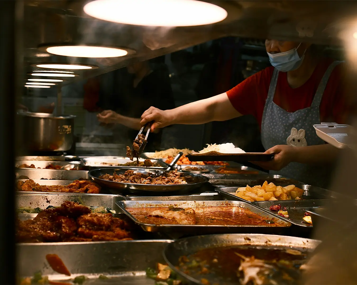 A woman choosing food from a tray at a food stall, carefully selecting dishes as part of the casual self-service dining experience.
