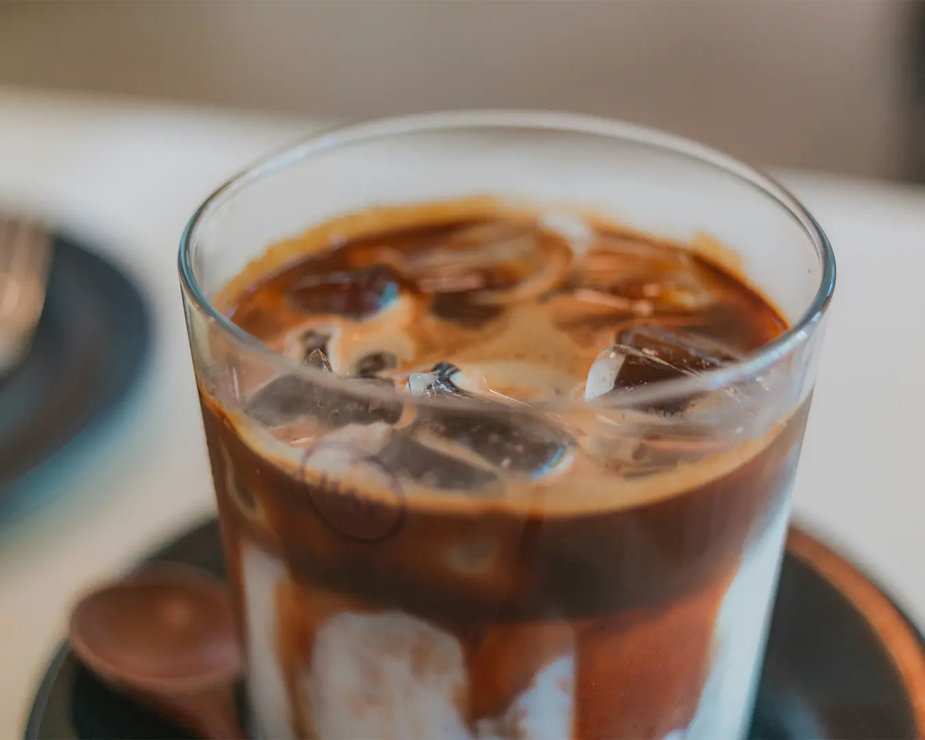 Close-up of an iced coffee in a glass cup on a saucer, featuring dark espresso swirls over creamy milk, evoking a refreshing and indulgent vibe.