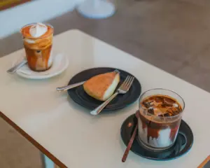 A cozy café table with two drinks: an iced coffee and a cappuccino with whipped cream, plus a pastry on a black plate. The scene feels warm and inviting.