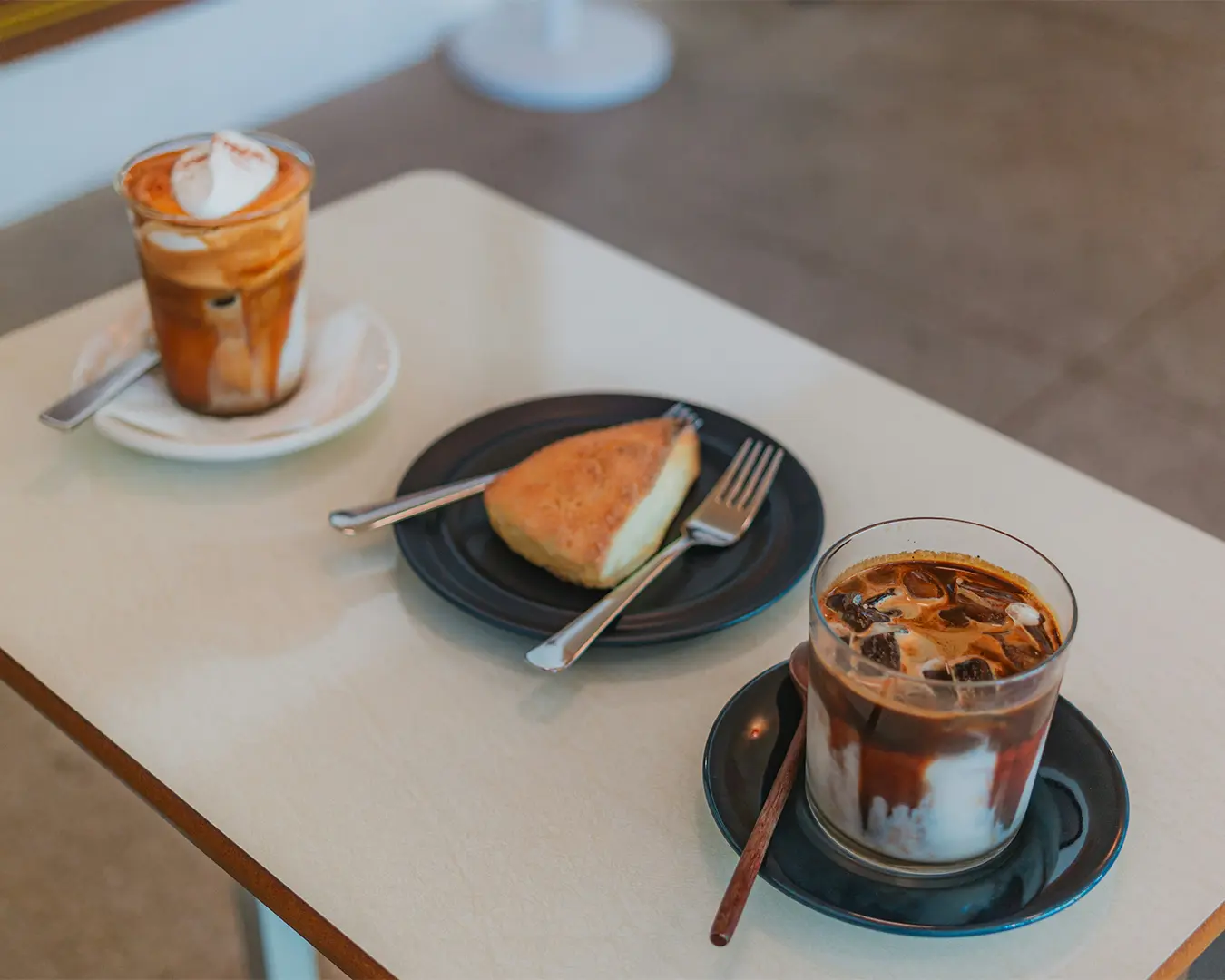 A cozy café table with two drinks: an iced coffee and a cappuccino with whipped cream, plus a pastry on a black plate. The scene feels warm and inviting.