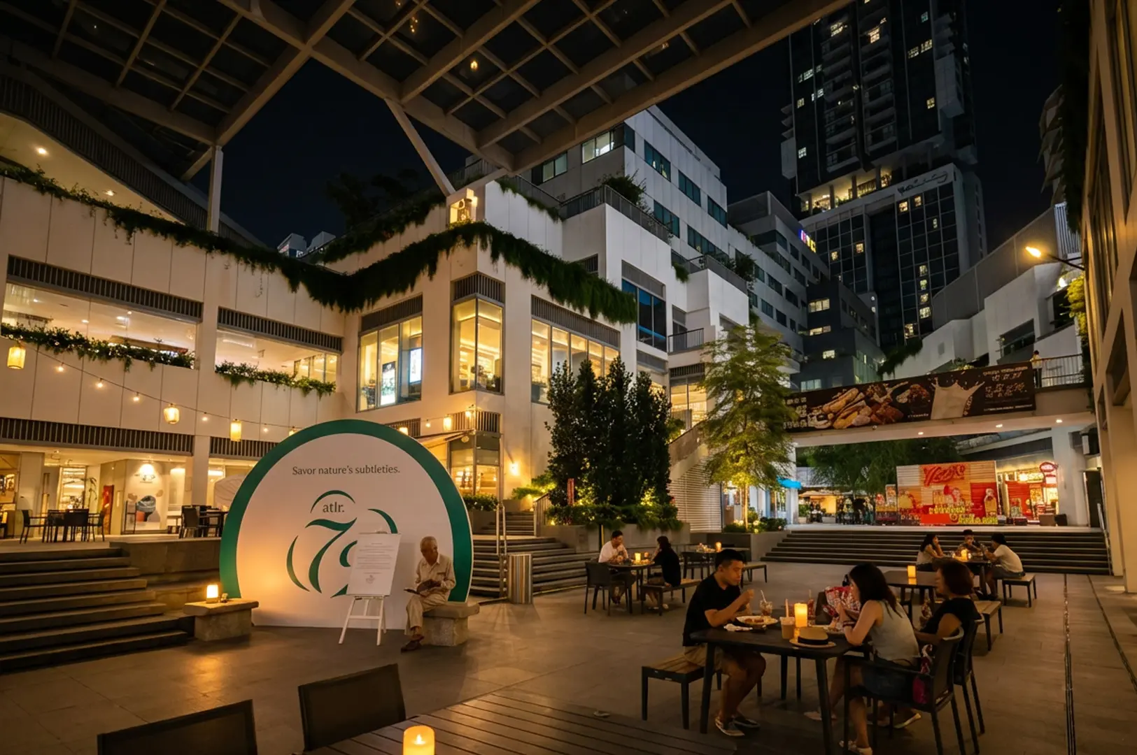 Modern outdoor dining plaza at night with people eating at tables, surrounded by white multi-level buildings with greenery and warm lighting